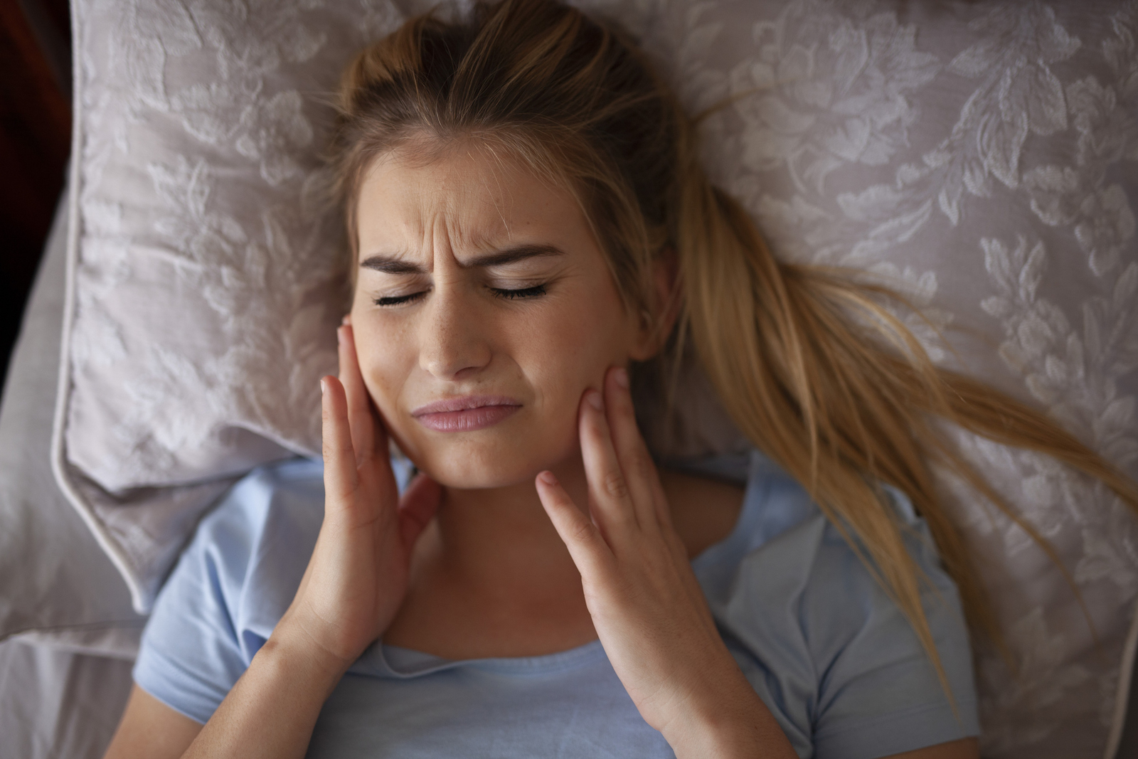 woman laying in bed feeling her sore jaw after grinding teeth