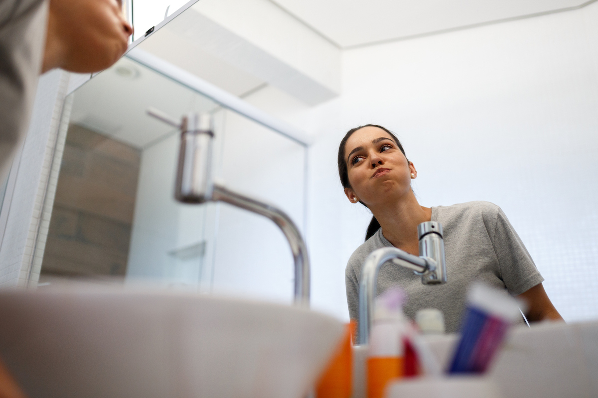 woman in bathroom standing over sink and looking in mirror while using mouthwash